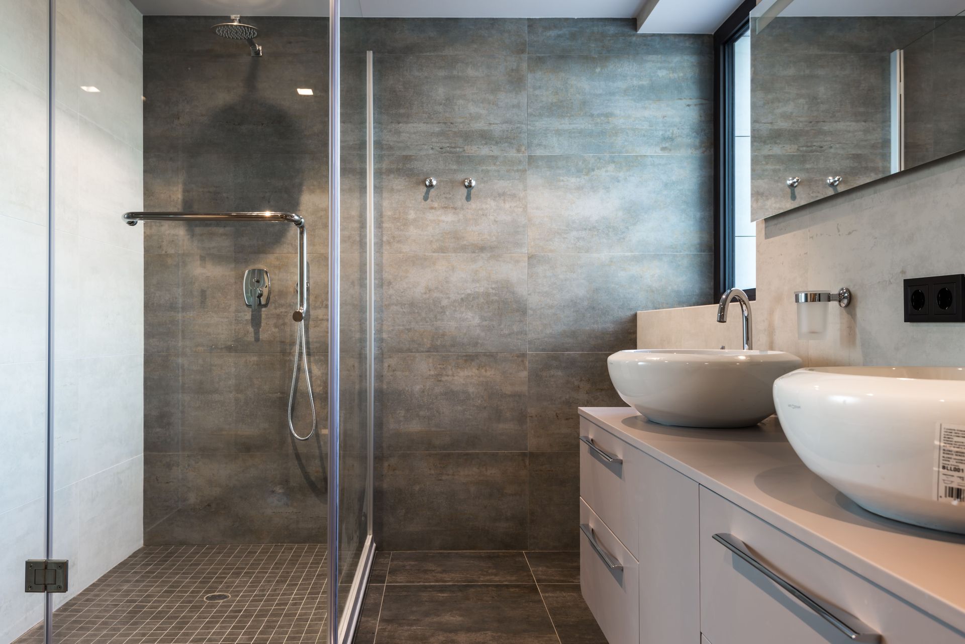 Salle de bains moderne avec douche en verre, murs carrelés gris et meuble-lavabo double avec vasques rondes.