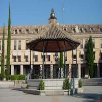 Mirador en una plaza con un edificio al fondo, con escalones de piedra y árboles altos bajo un cielo azul.