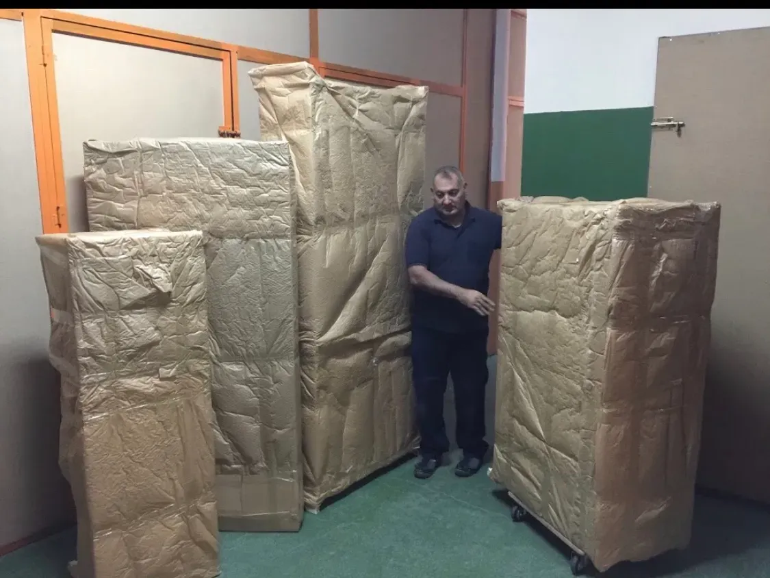 Un hombre de pie entre grandes paquetes envueltos en cartón en un pasillo de un almacén.