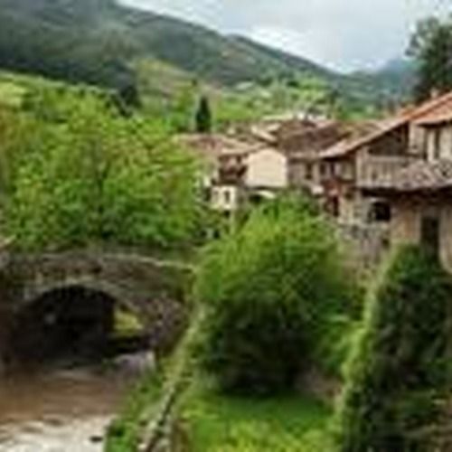 Puente de piedra sobre un río con casas de un pueblo y montañas al fondo.