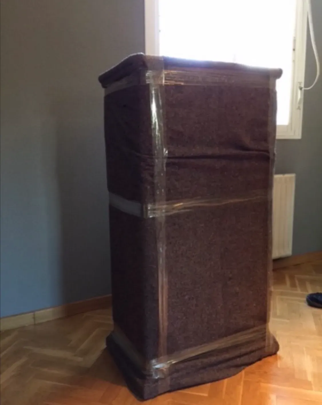 Un mueble  envuelto con manta marrón se encuentra junto a una ventana sobre un suelo de madera.