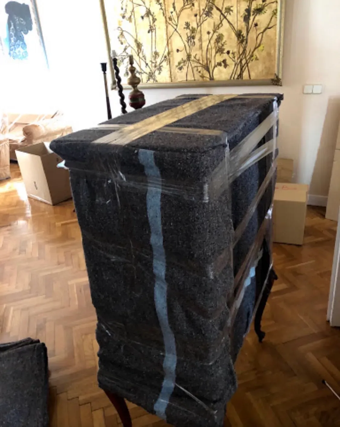 Muebles verticales envueltos en tela negra en una habitación soleada con suelo de parqué y decoración floral en la pared.