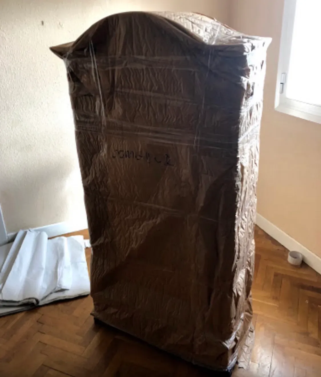 Armario vertical envuelto en plástico en una habitación cerca de una ventana y con suelo de madera.