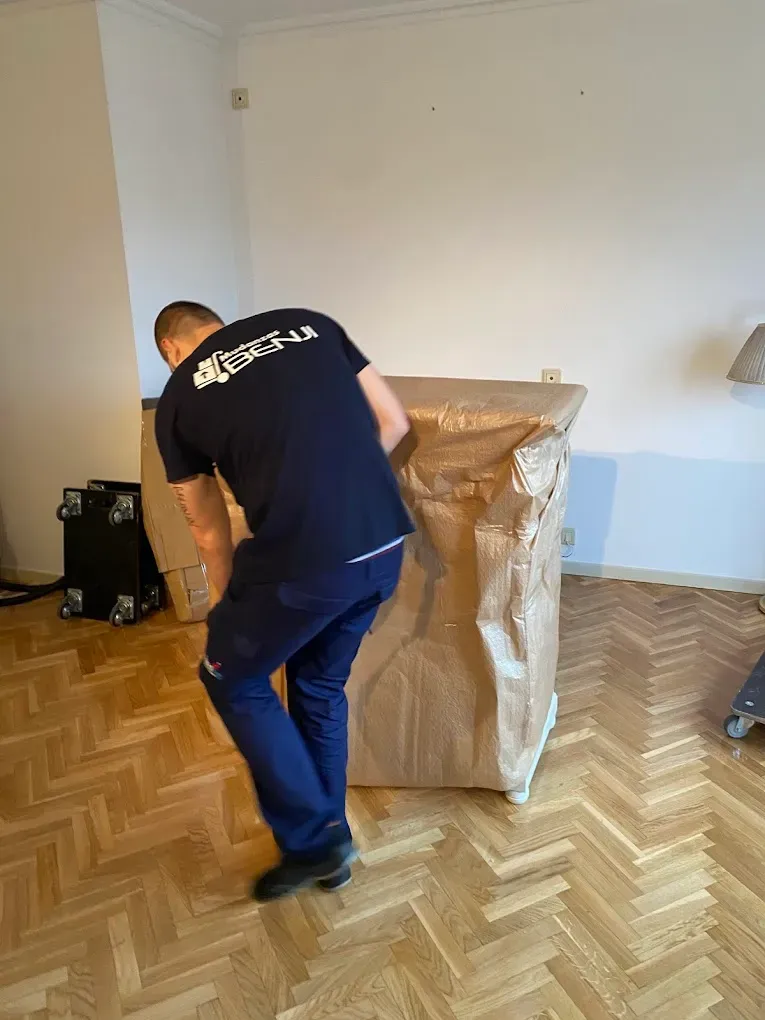 Persona trasladando un objeto grande envuelto en una habitación vacía con suelo de parqué.