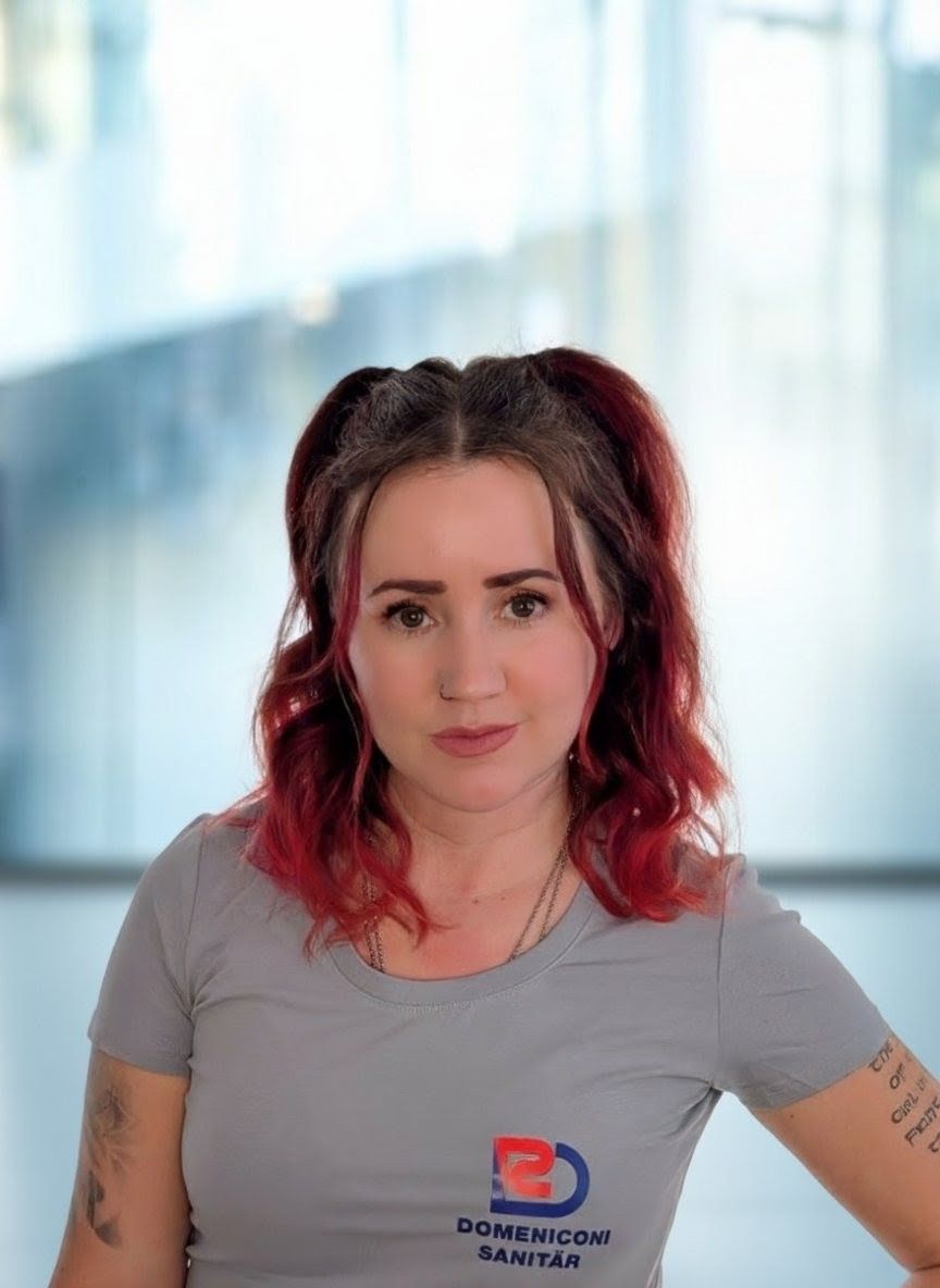 Frau in grauem Hemd mit Logo, lächelnd, in einem Innenraum.