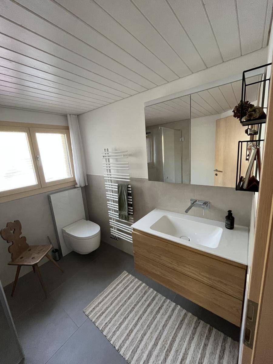 Badezimmer mit Waschtisch aus Holz, weissem Waschbecken, grossem Spiegel und Handtuchwärmer. Grauer Boden, gestreifter Teppich.