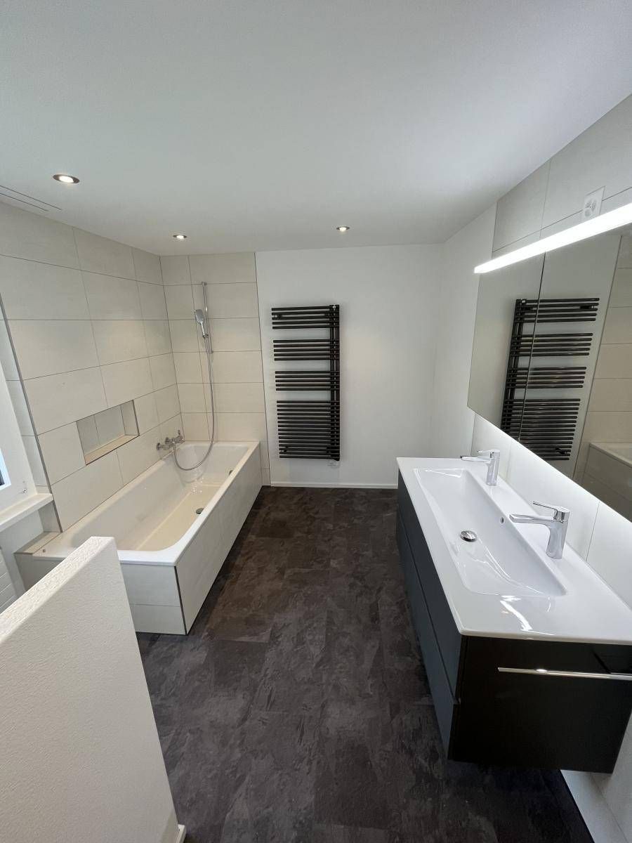 Modernes Badezimmer mit weisser Badewanne, dunkelgrauem Boden, dunkelgrauem Schrank und schwarzem Handtuchwärmer.