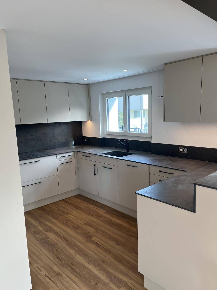 Moderne, helle Küche mit hellgrauen Schränken, dunklen Arbeitsplatten und Holzbodenoptik.