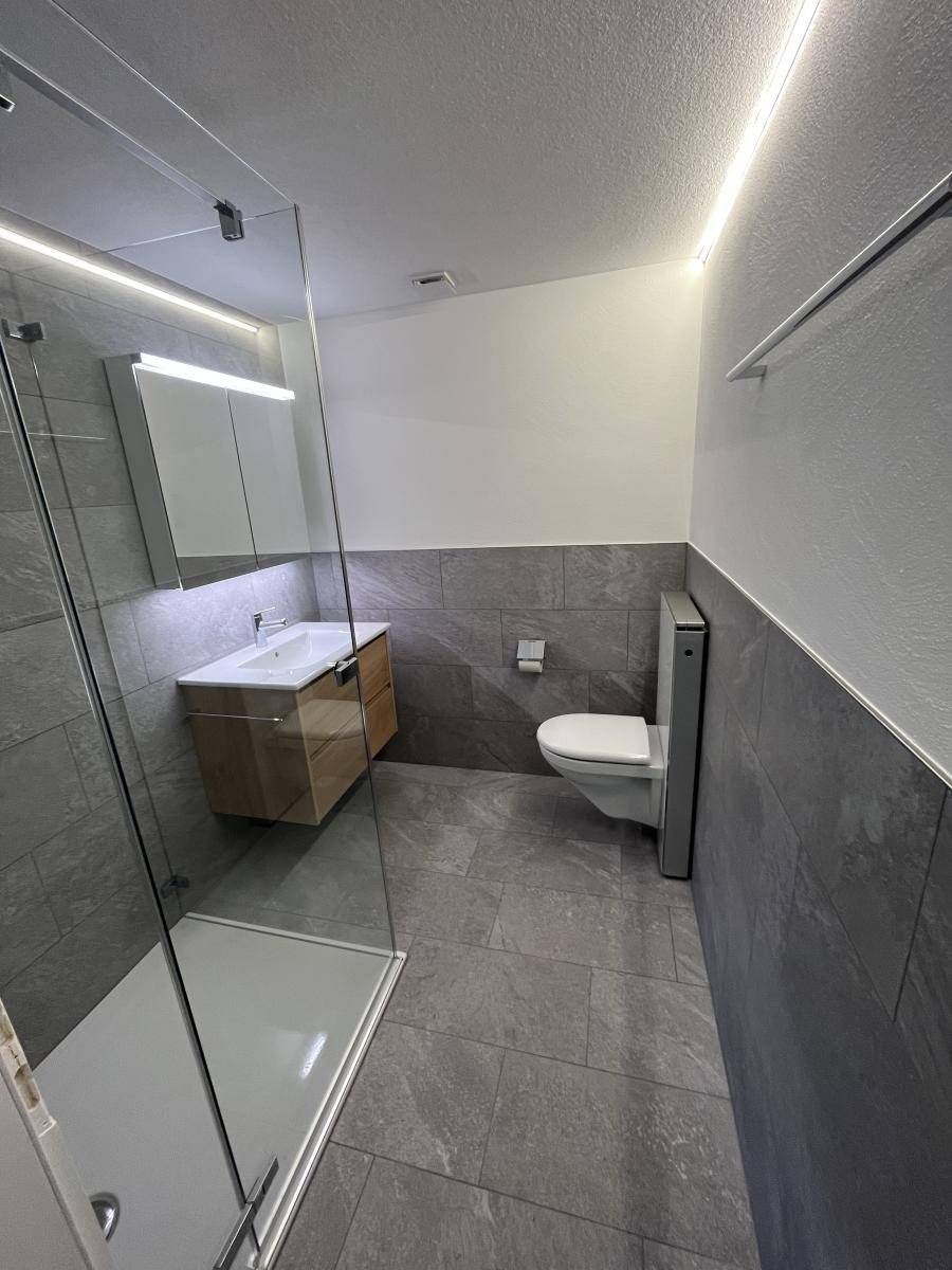 Modernes Badezimmer mit Glasdusche, schwebendem Waschtisch, grauen Fliesen und Toilette.