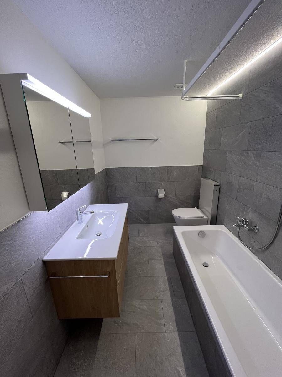 Modernes Badezimmer mit grau gefliesten Wänden, einem Waschtisch aus Holz und einer Badewanne.