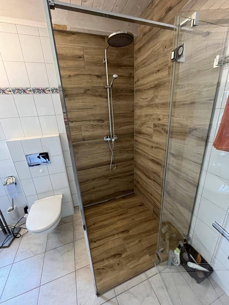 Badezimmer mit Glasdusche, Wandfliesen in Holzoptik, weisser Toilette und weissem Fliesenboden.