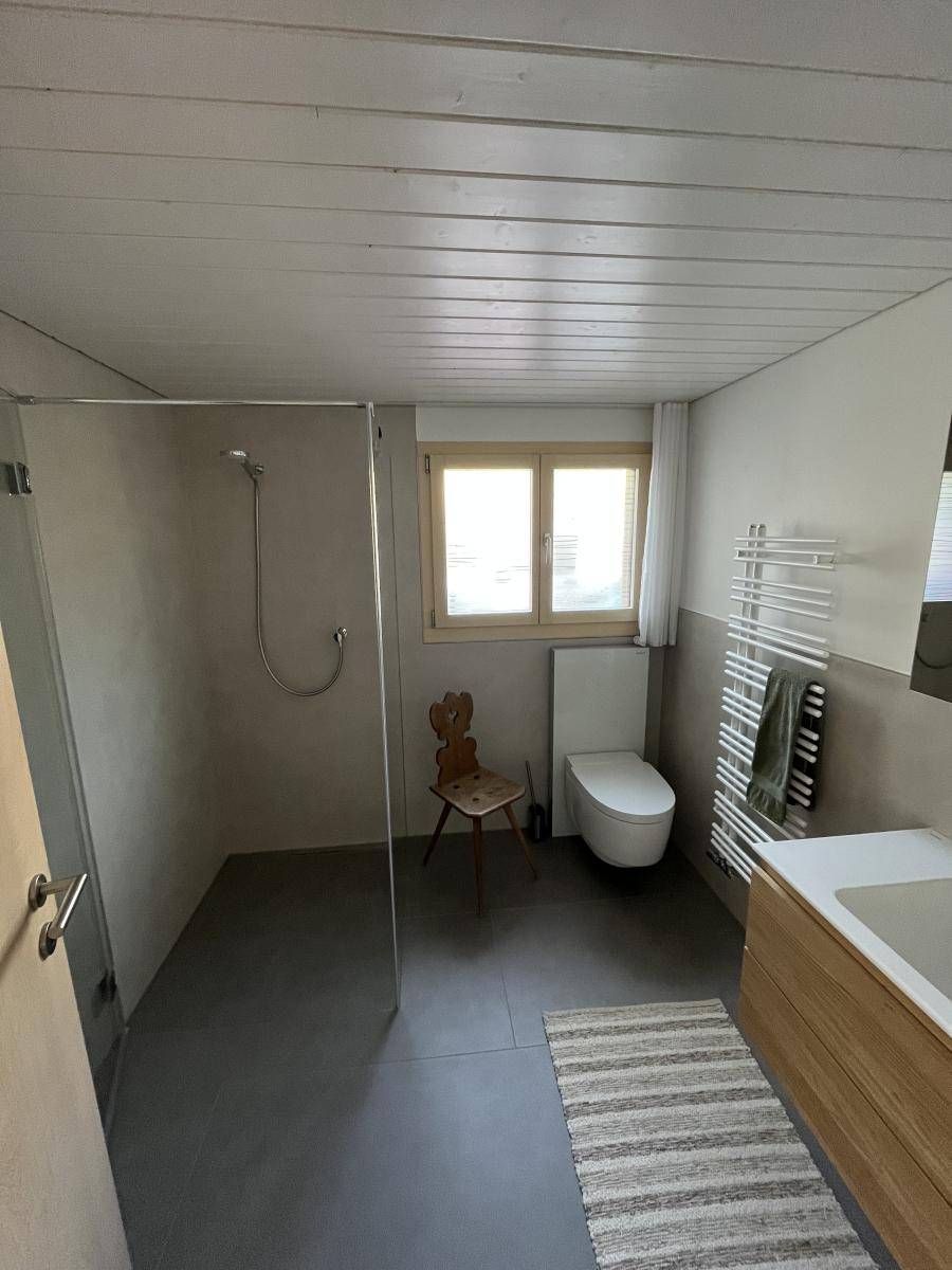 Badezimmer mit Dusche, WC, Fenster, Waschtisch aus Holz und grauem Boden.