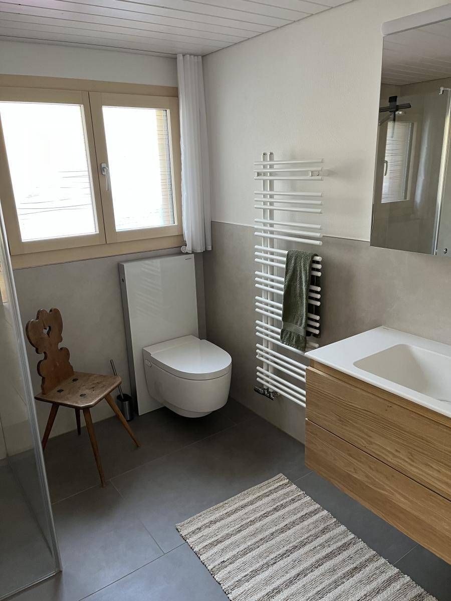 Badezimmer mit Toilette, Waschtisch aus Holz, Fenster, Handtuchhalter und dekorativem Stuhl. Farbgestaltung in Grau und Weiss.