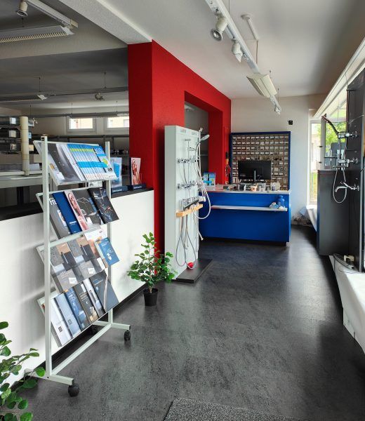 Innenansicht eines Ausstellungsraums mit Badezimmerausstattung. Rote Säule, blauer Tresen, grauer Boden, Produktauslagen und Zeitschriften.