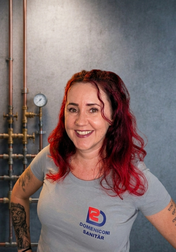 Frau in grauem Hemd mit Logo, lächelnd, in einem Innenraum.