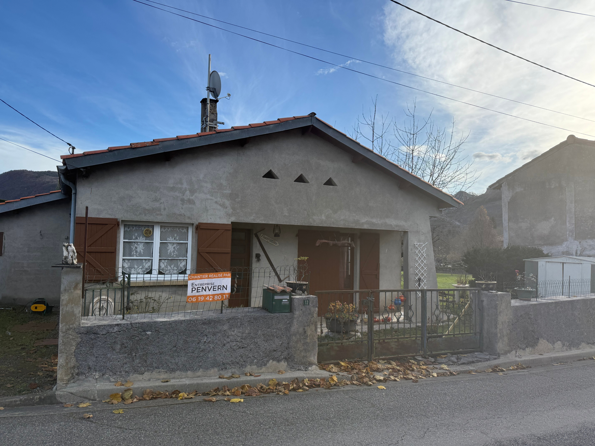 Petite maison grise avec volets et porte marron, panneau « À vendre » devant.