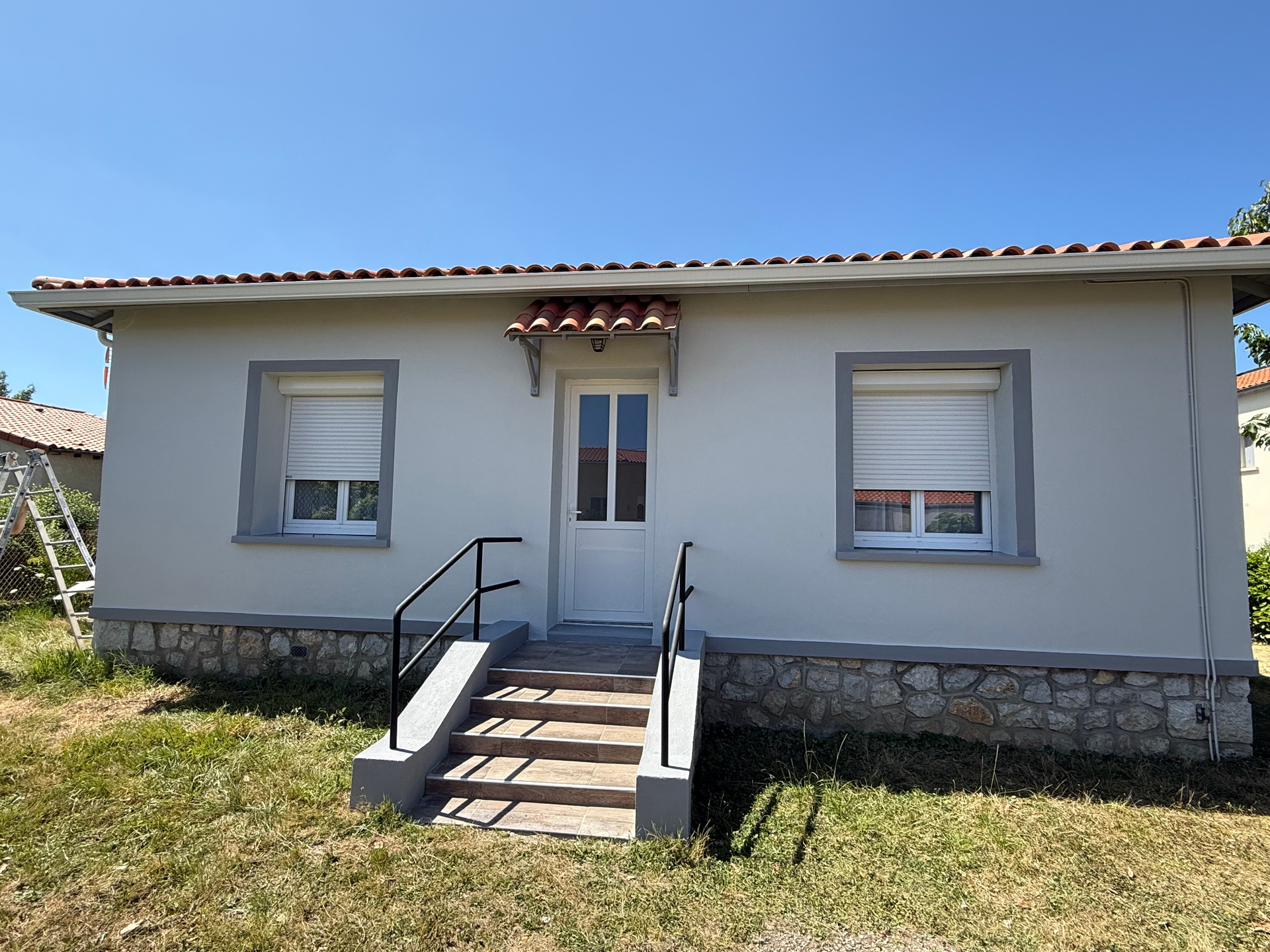 Petite maison gris clair avec soubassement en pierre, marches menant à la porte blanche.