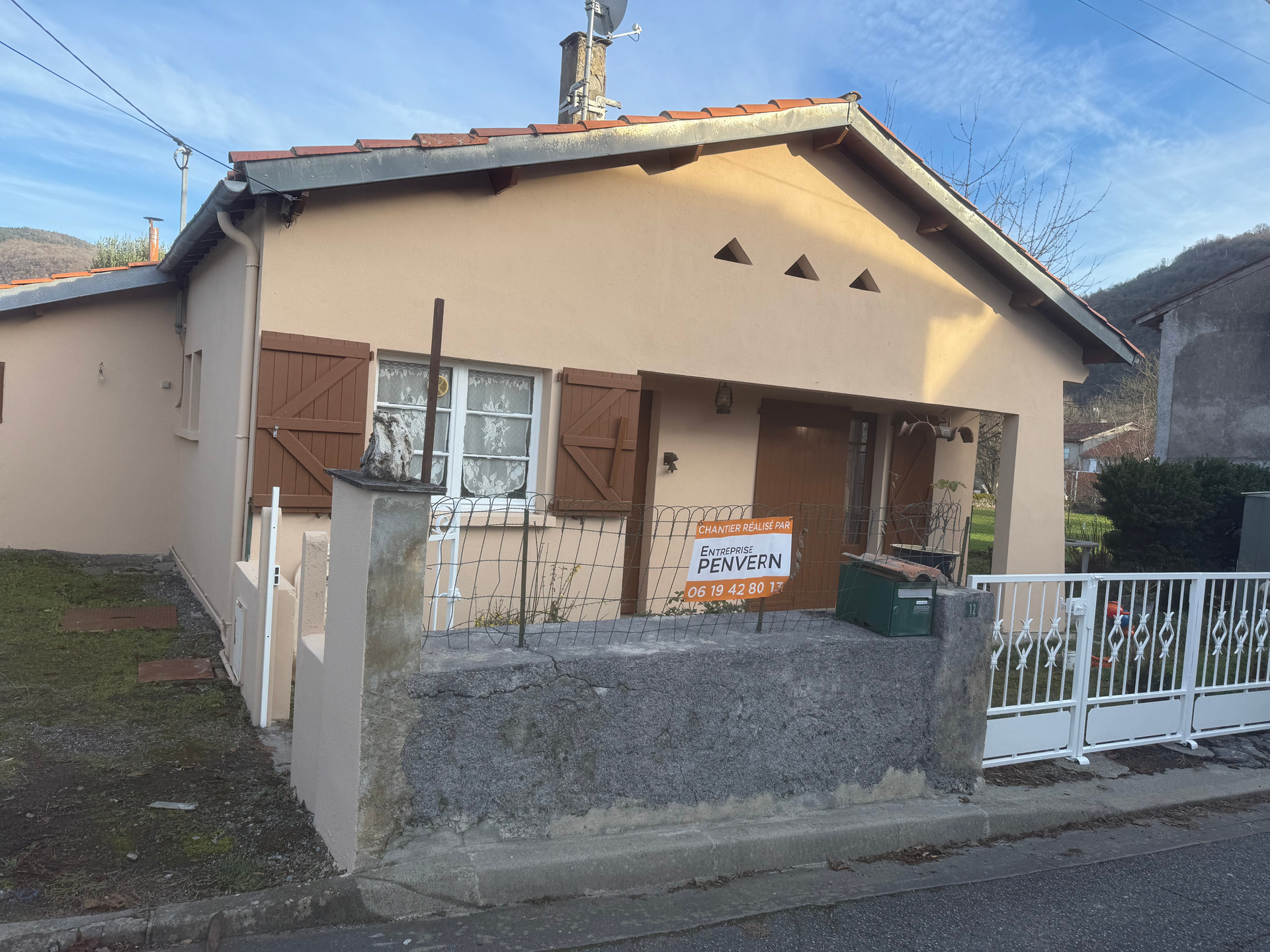 Maison couleur sable avec des volets marron, un mur en béton et un portail blanc.