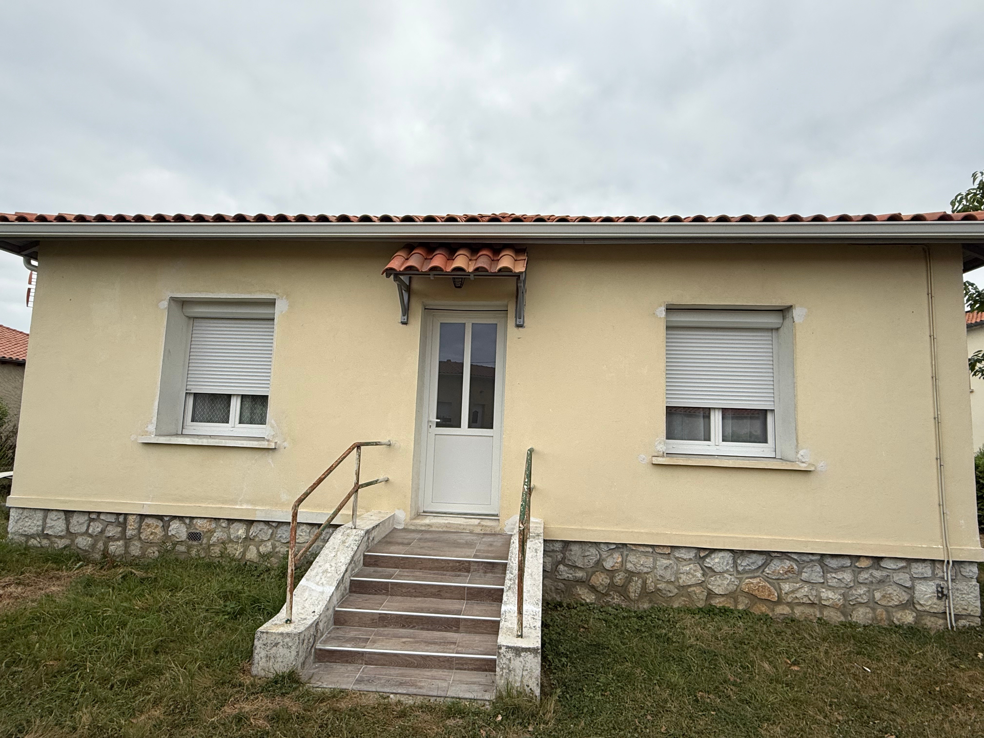 Maison beige avec porte blanche, deux fenêtres à stores, fondations en pierre et marches.