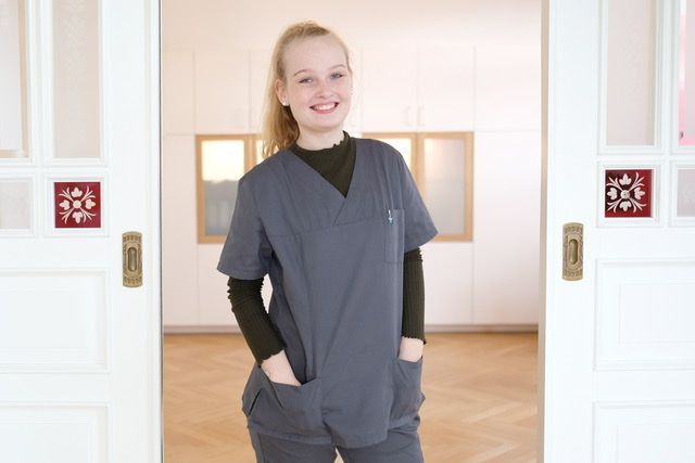 Eine Frau im OP-Kittel steht mit den Händen in den Taschen in einem Türrahmen.