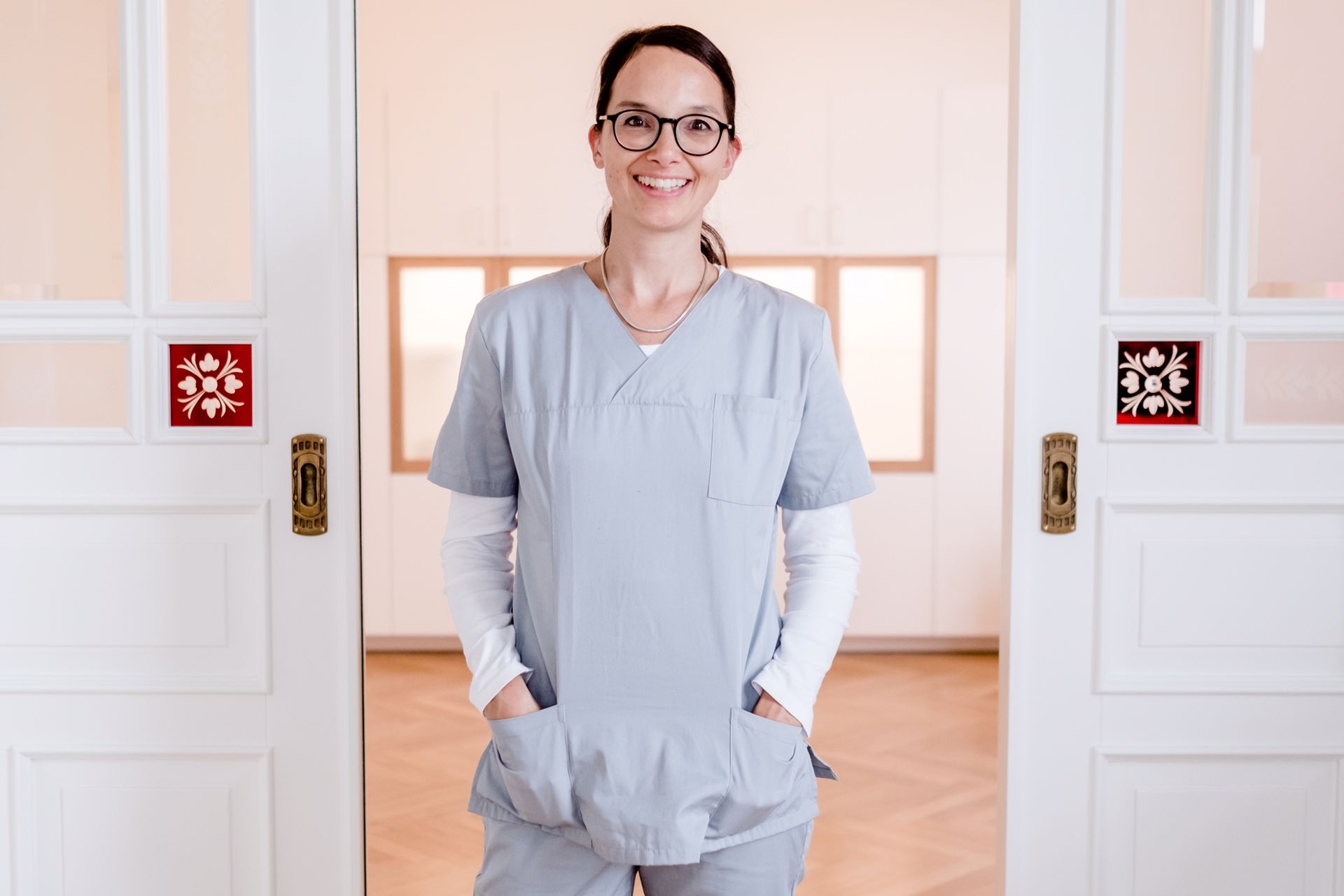 Eine Krankenschwester steht mit den Händen in den Taschen vor einer Tür.