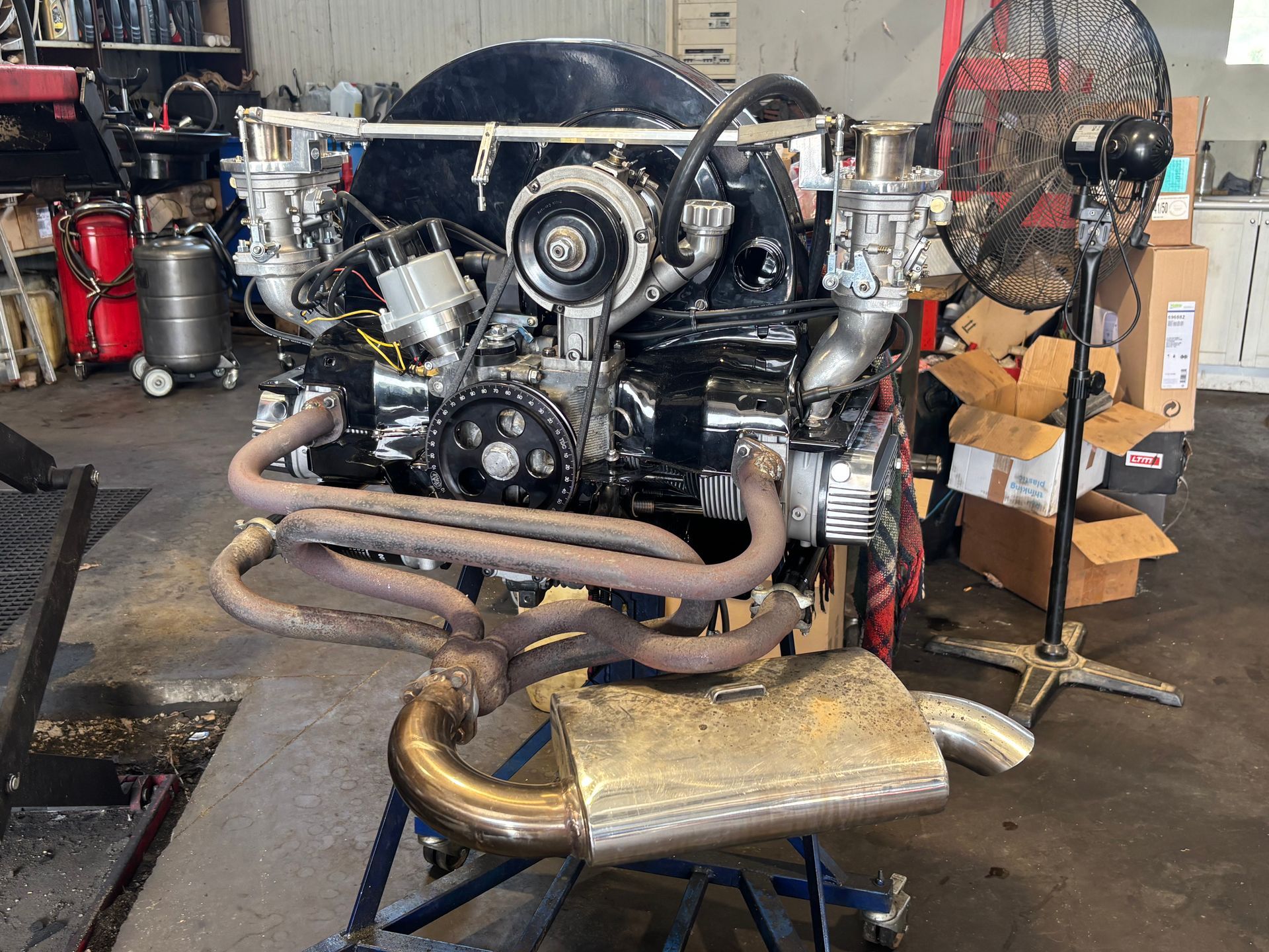 Assemblage d'un moteur sur le sol d'un atelier, avec tuyauterie d'échappement apparente et ventilateur sur pied en arrière-plan.
