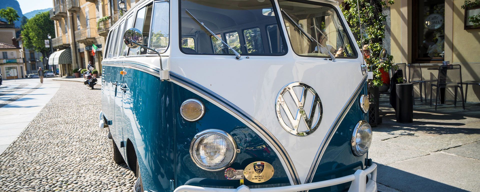 Une camionnette Volkswagen vintage bleue et blanche est garée dans une rue pavée d'une ville ensoleillée.