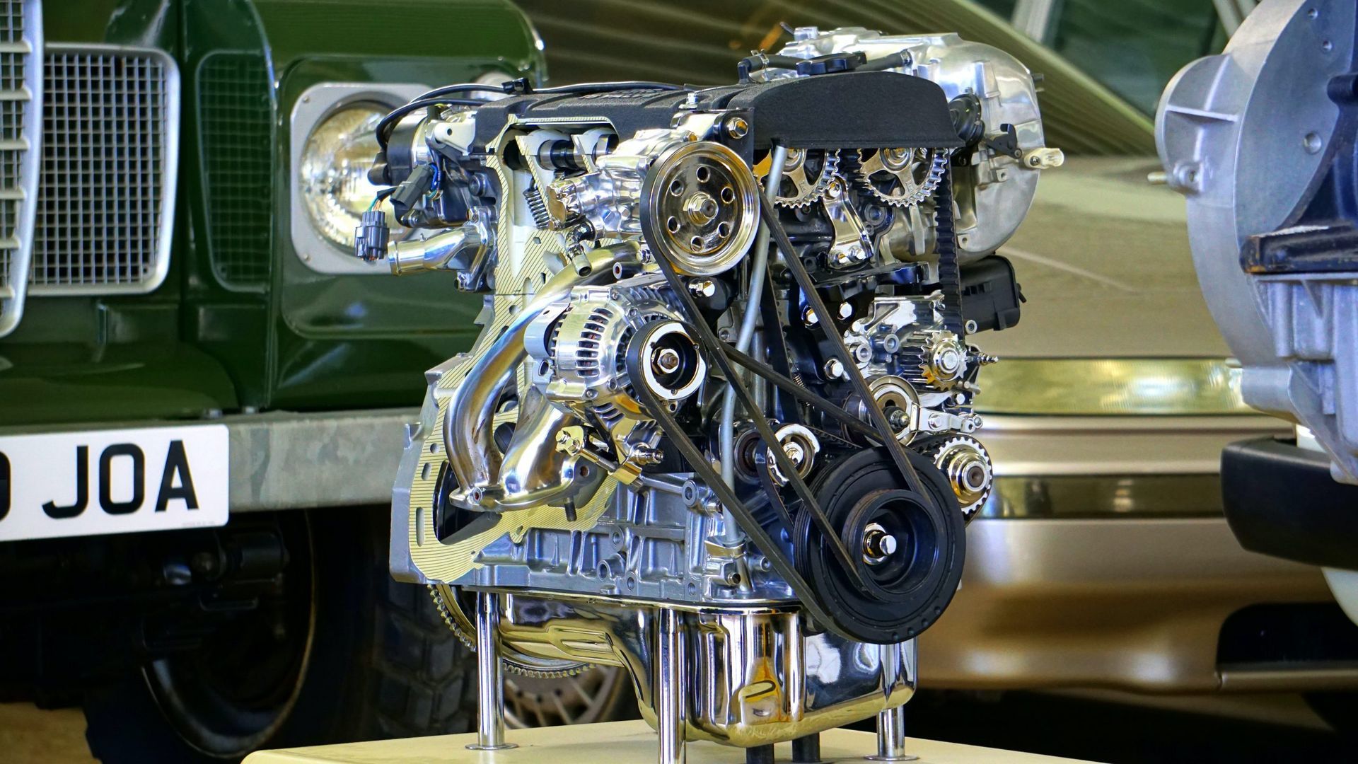 Gros plan sur un moteur de voiture poli exposé, avec un véhicule ancien vert en arrière-plan.