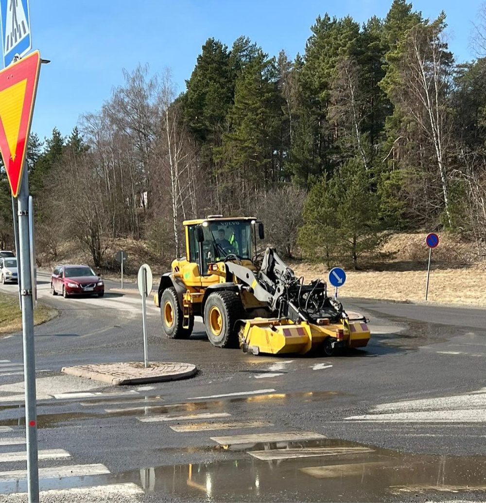 Keltainen tiehöylä tasoittaa märkää tietä liikenneympyrässä. Taustalla näkyy auto ja puita.