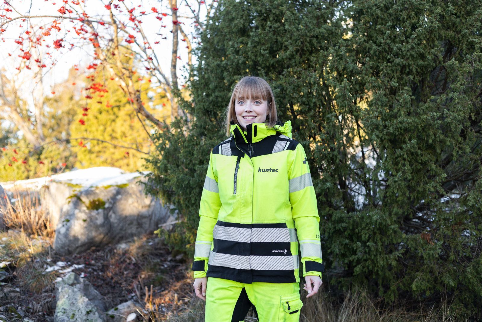 Henkilö kirkkaan keltaisessa huomiotakissa seisomassa ulkona puiden ja kivien lähellä.