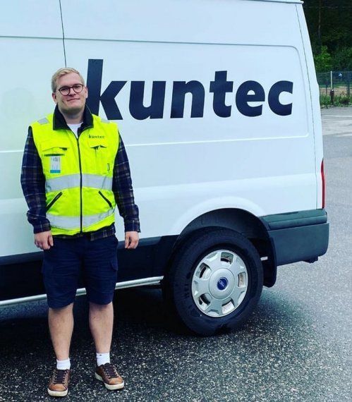 Henkilö keltaisessa huomioliivissä seisoo valkoisen Kuntec-pakettiauton vieressä märällä kadulla.