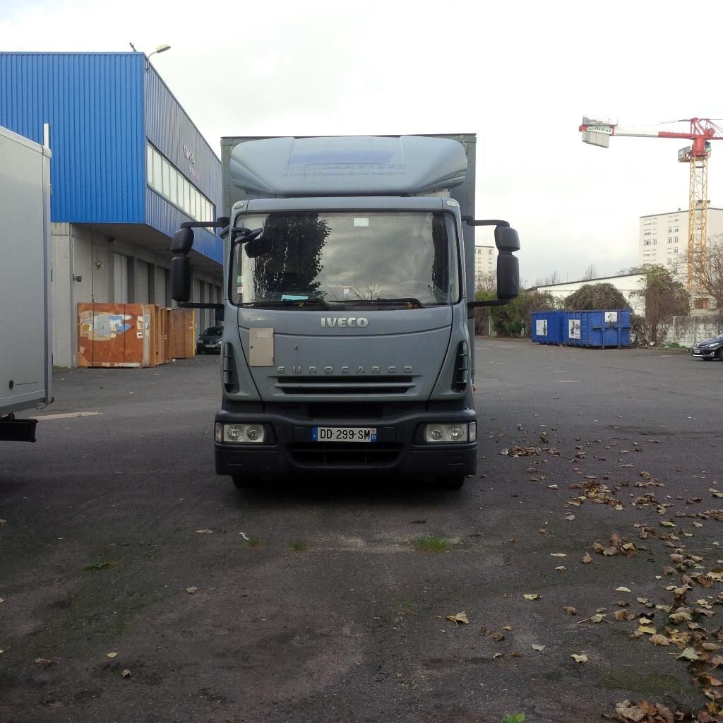 Camion gris stationné sur un terrain industriel à côté d'entrepôts bleus et de bennes à ordures.