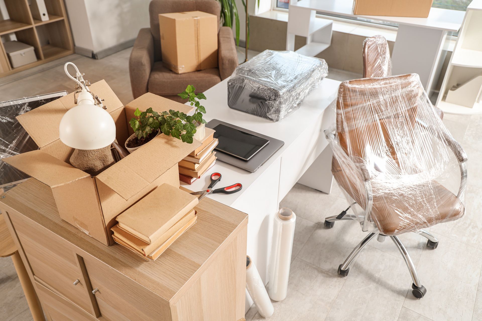 Des cartons de déménagement et une chaise emballée dans un bureau lumineux avec bureau et lampe.
