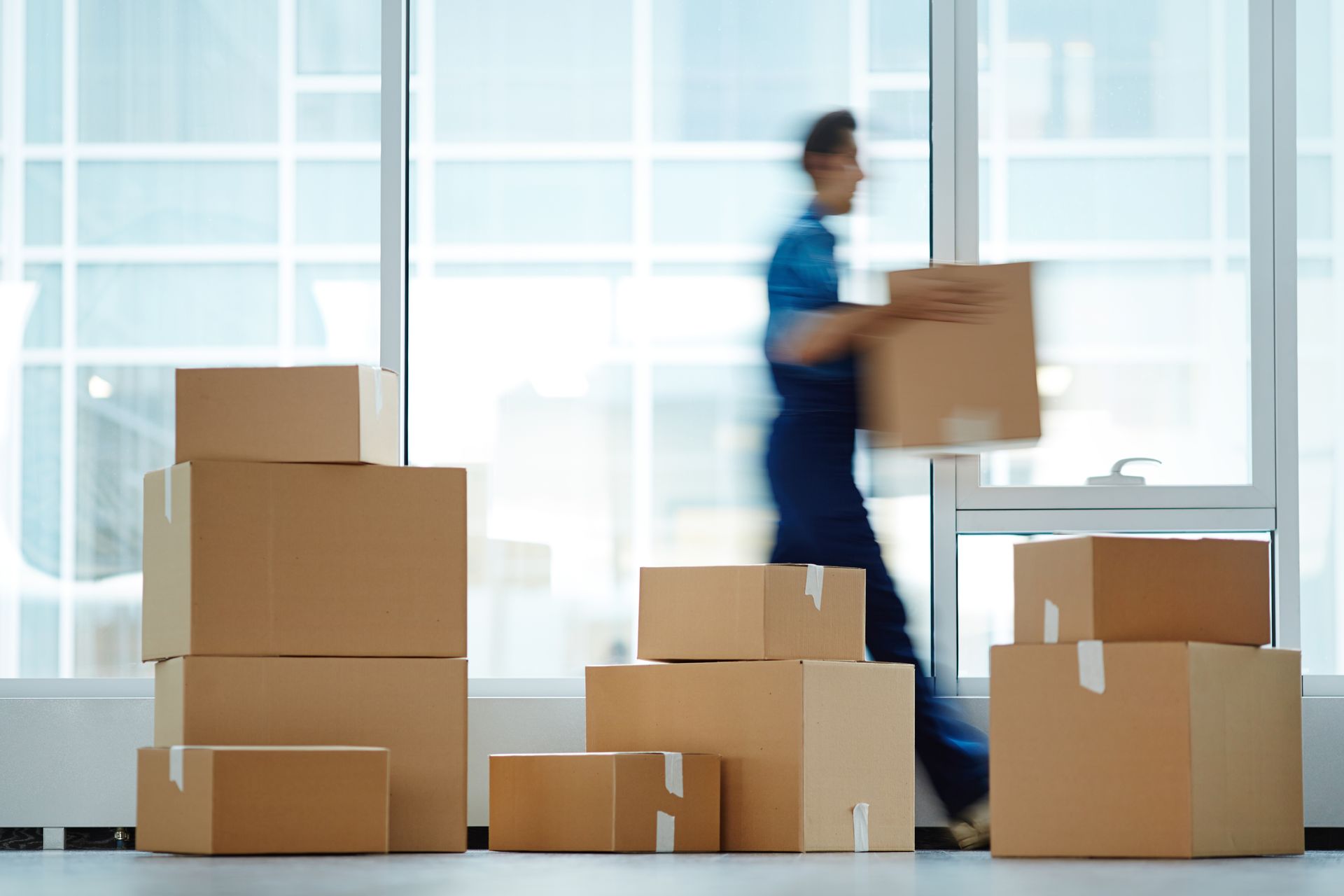 Une personne transporte un carton parmi des cartons de déménagement empilés dans un bureau lumineux.