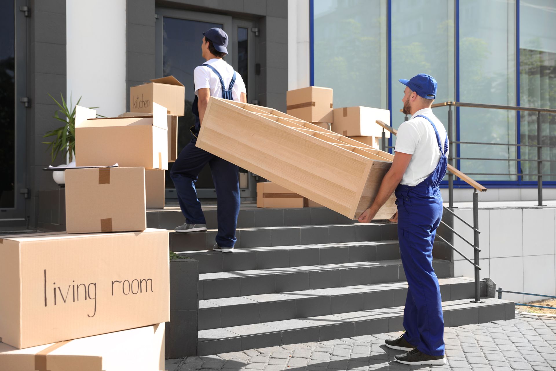 Deux déménageurs transportent un long carton en haut des marches d'un immeuble, des cartons de déménagement empilés à proximité.