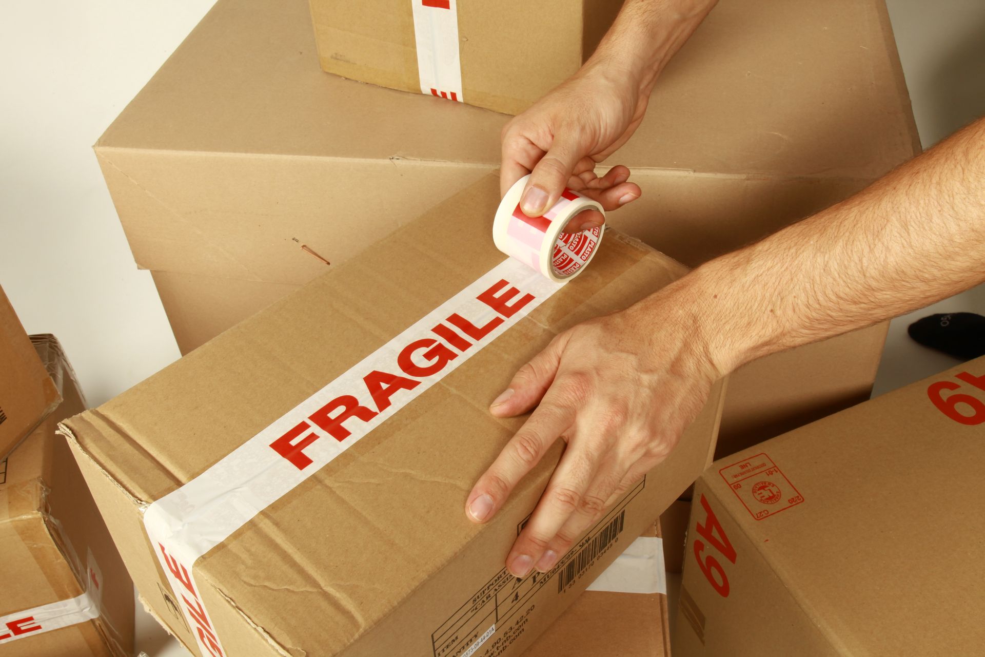 Des mains scellent un carton avec du ruban adhésif rouge portant la mention FRAGILE parmi des cartons de déménagement empilés.