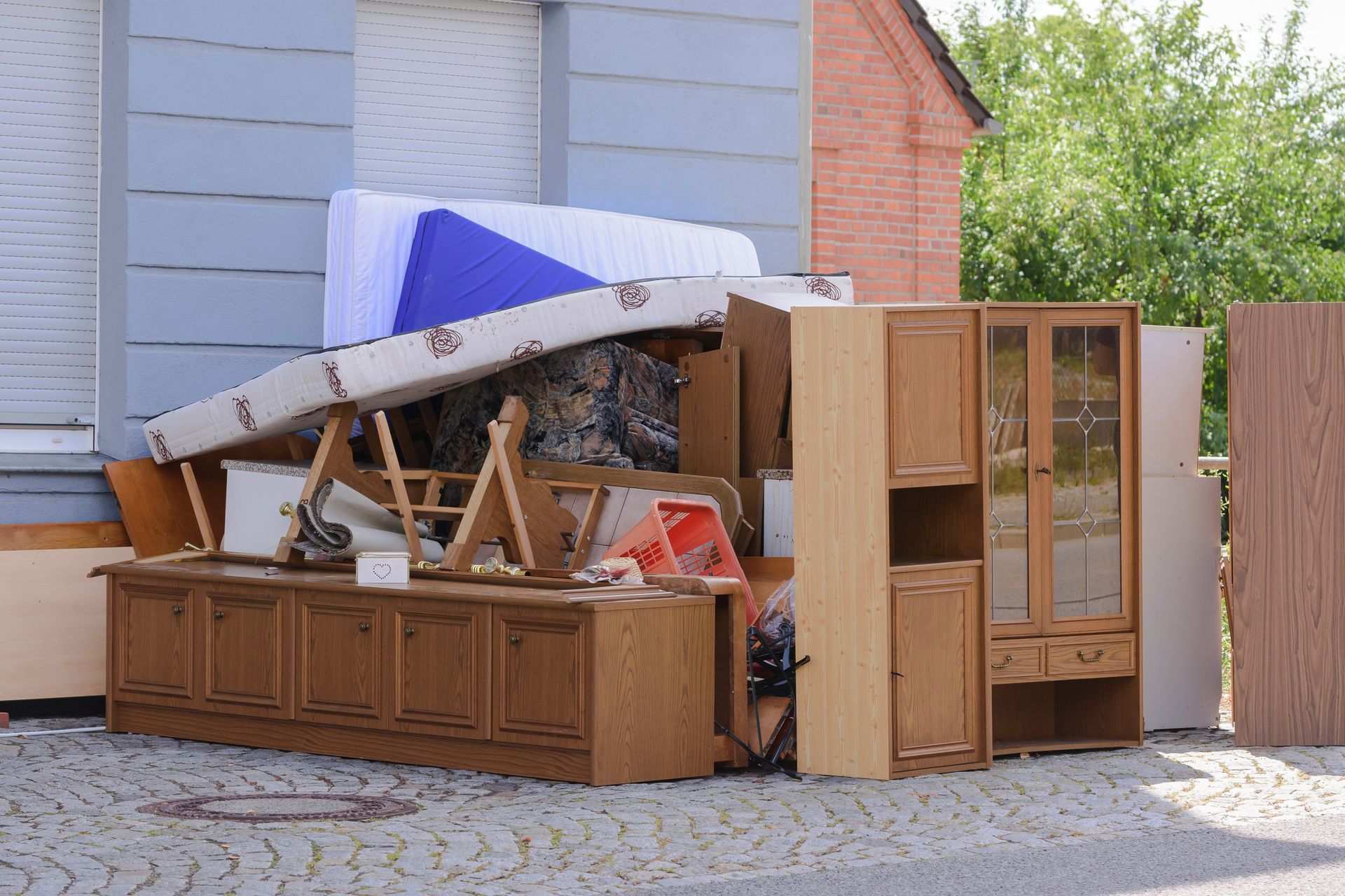 Un tas de meubles et de débris jetés, comprenant des armoires et un matelas, s'entasse à côté d'une maison.