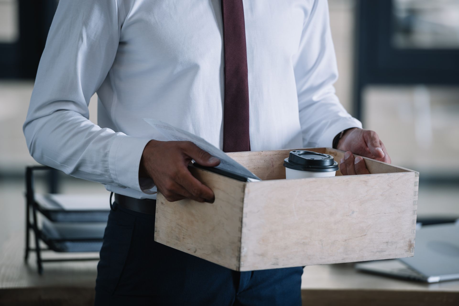 Une personne en chemise blanche et cravate tient une boîte en carton contenant des fournitures de bureau dans un environnement de bureau.