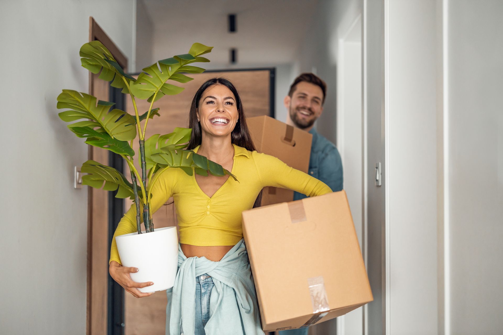 Un couple souriant transportant des cartons et une plante en pot dans un couloir, emménageant dans une nouvelle maison