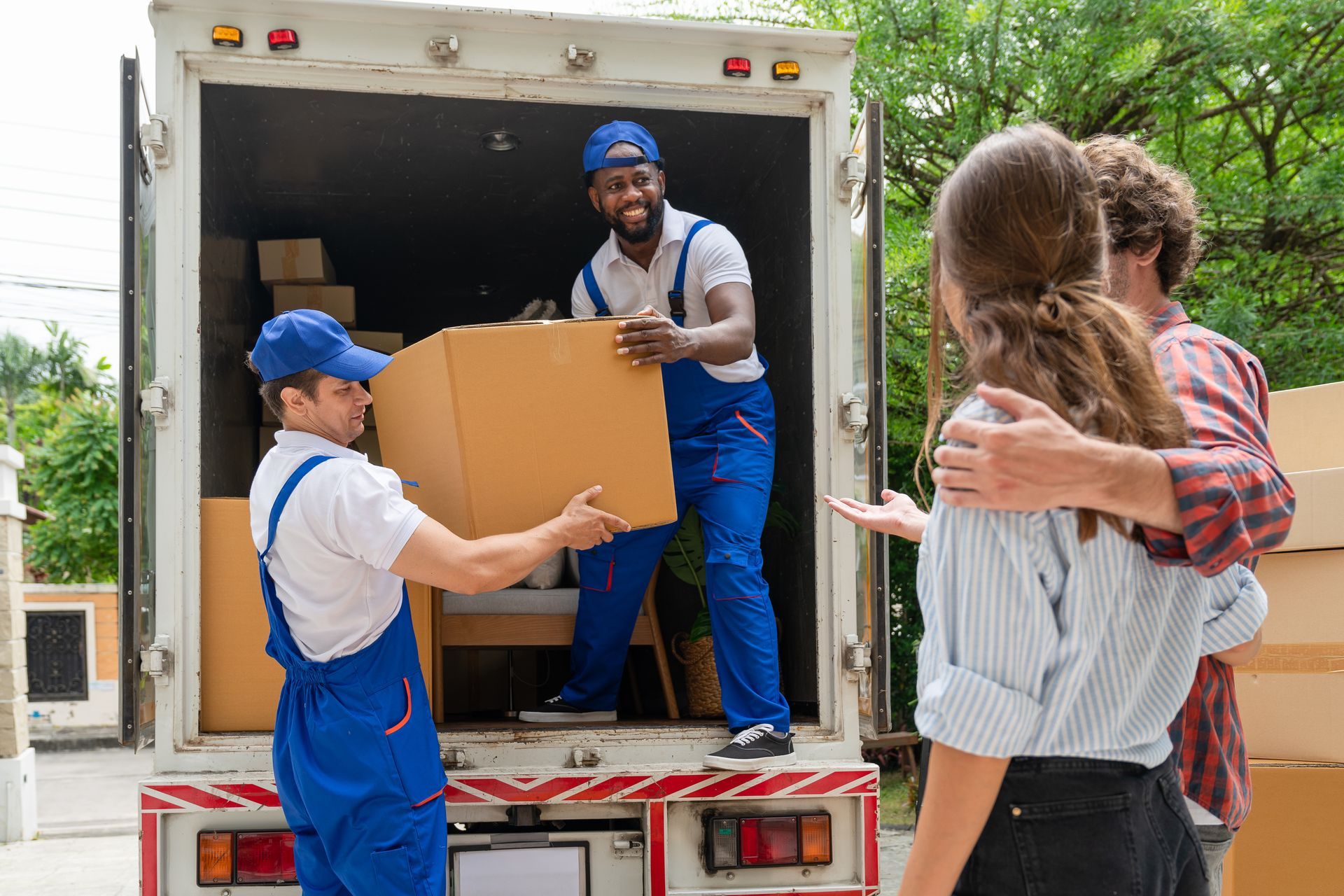 Des déménageurs déchargent des cartons d'un camion de déménagement tandis qu'un couple attend à proximité.