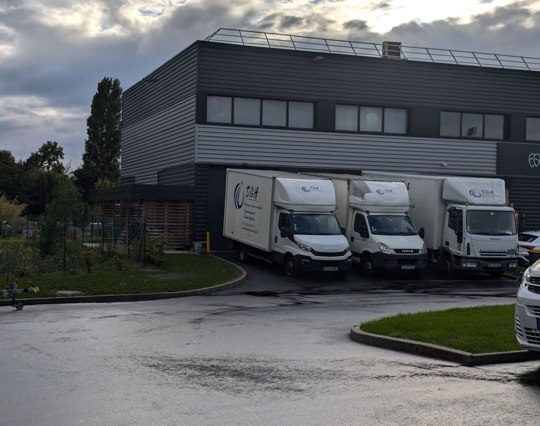 Trois camionnettes de livraison blanches garées à côté d'un bâtiment industriel gris sous un ciel nuageux.