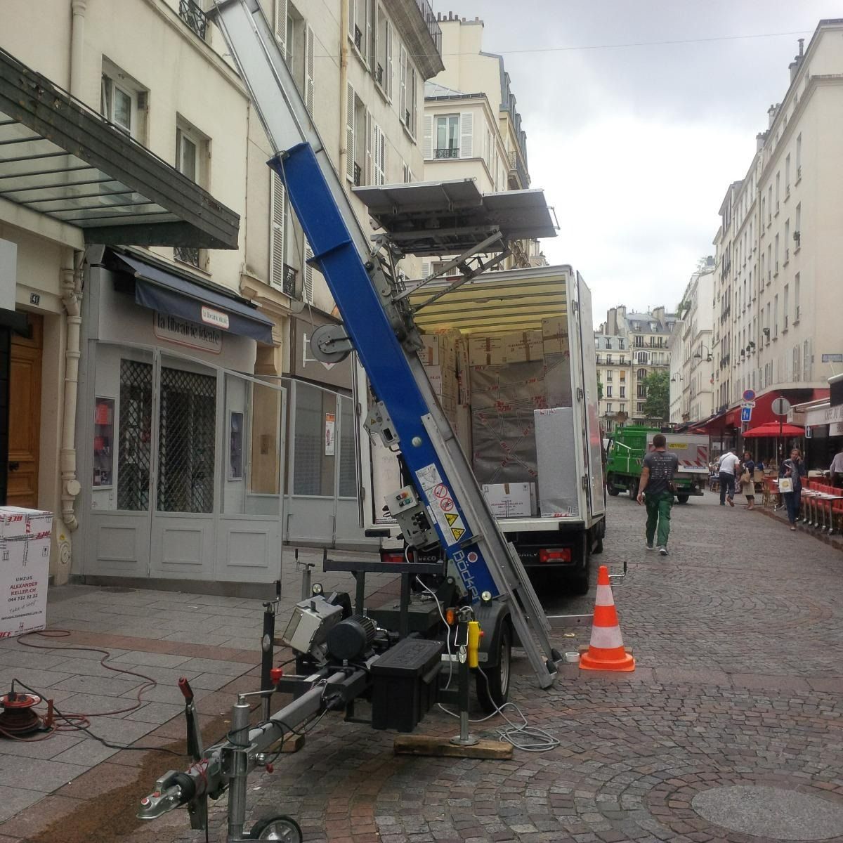 Une nacelle élévatrice bleue se trouve à côté d'un camion dans une rue pavée d'une ruelle de la ville.