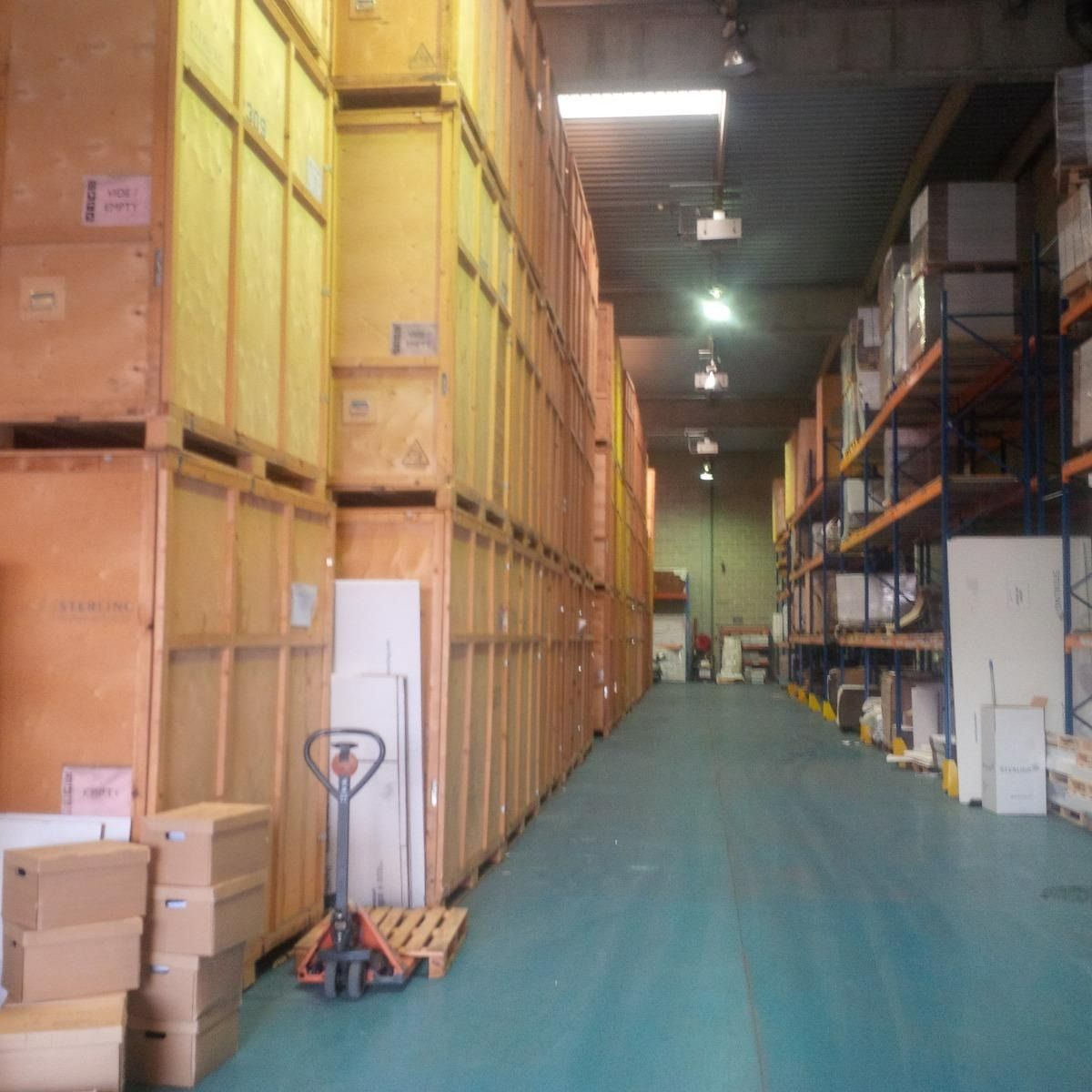 Une allée d'entrepôt avec des cartons empilés à gauche, des étagères à droite et un sol bleu.