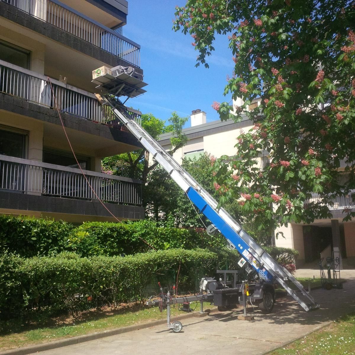 Une nacelle élévatrice bleue s'étend jusqu'au balcon d'un immeuble d'appartements donnant sur une cour intérieure arborée.