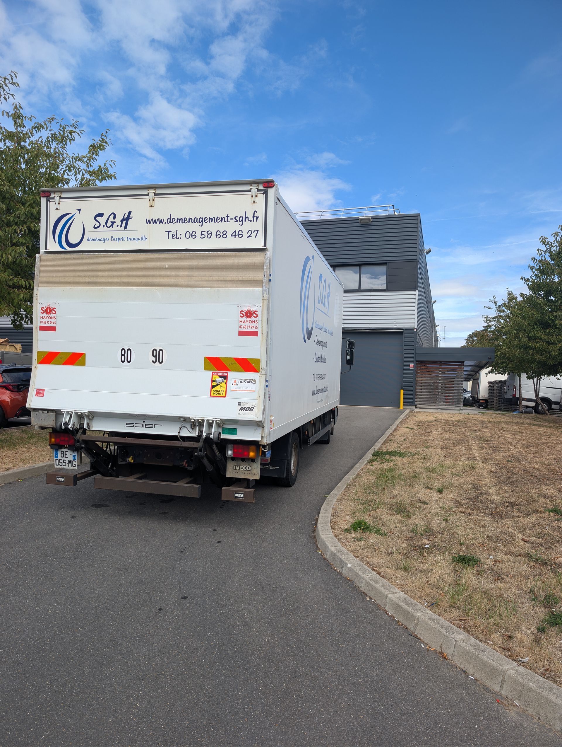 Un camion utilitaire blanc est garé dans une allée courbe à côté d'une maison moderne, sous un ciel bleu.