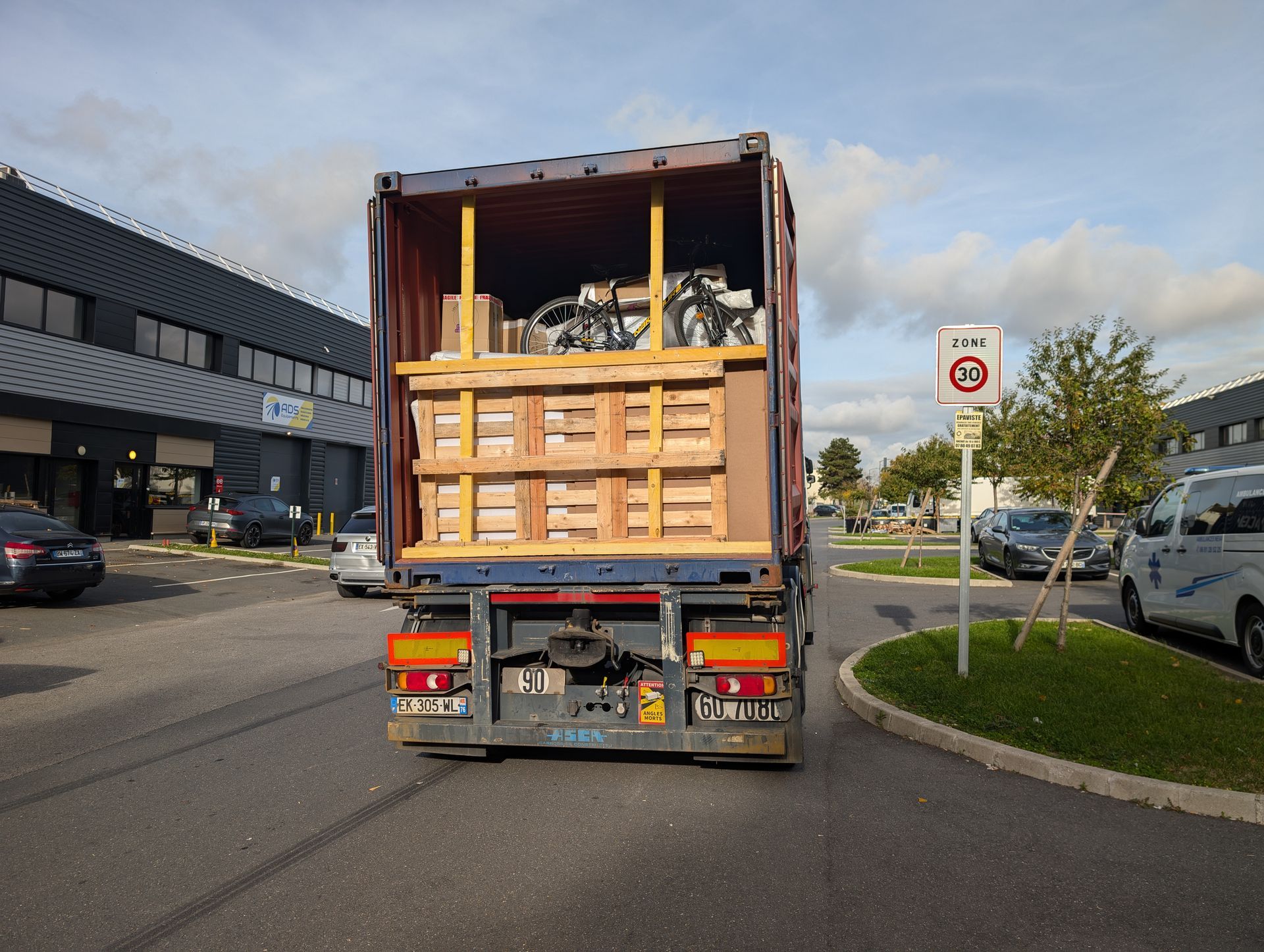Un camion chargé de palettes en bois circule en bord de route près d'un parking.