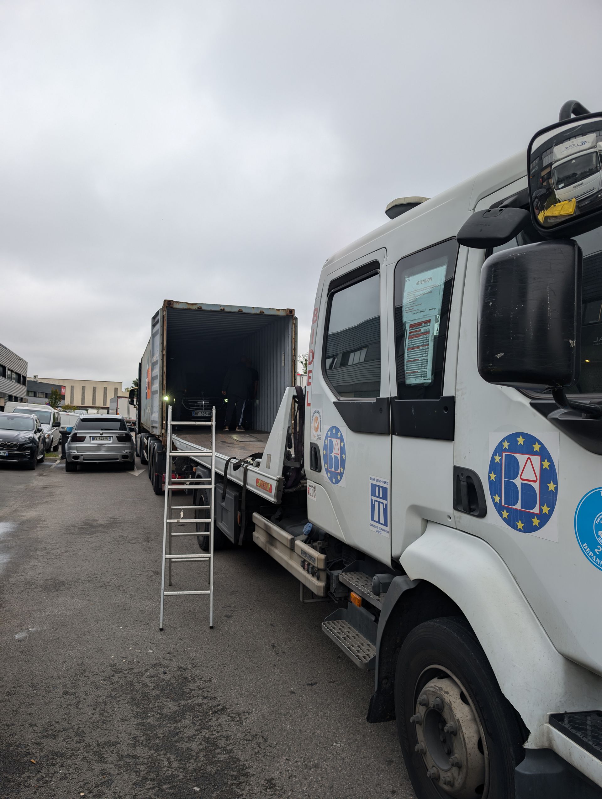 Camionnette de service blanche avec espace de chargement arrière ouvert et échelle, stationnée dans une rue mouillée sous un ciel nuageux.