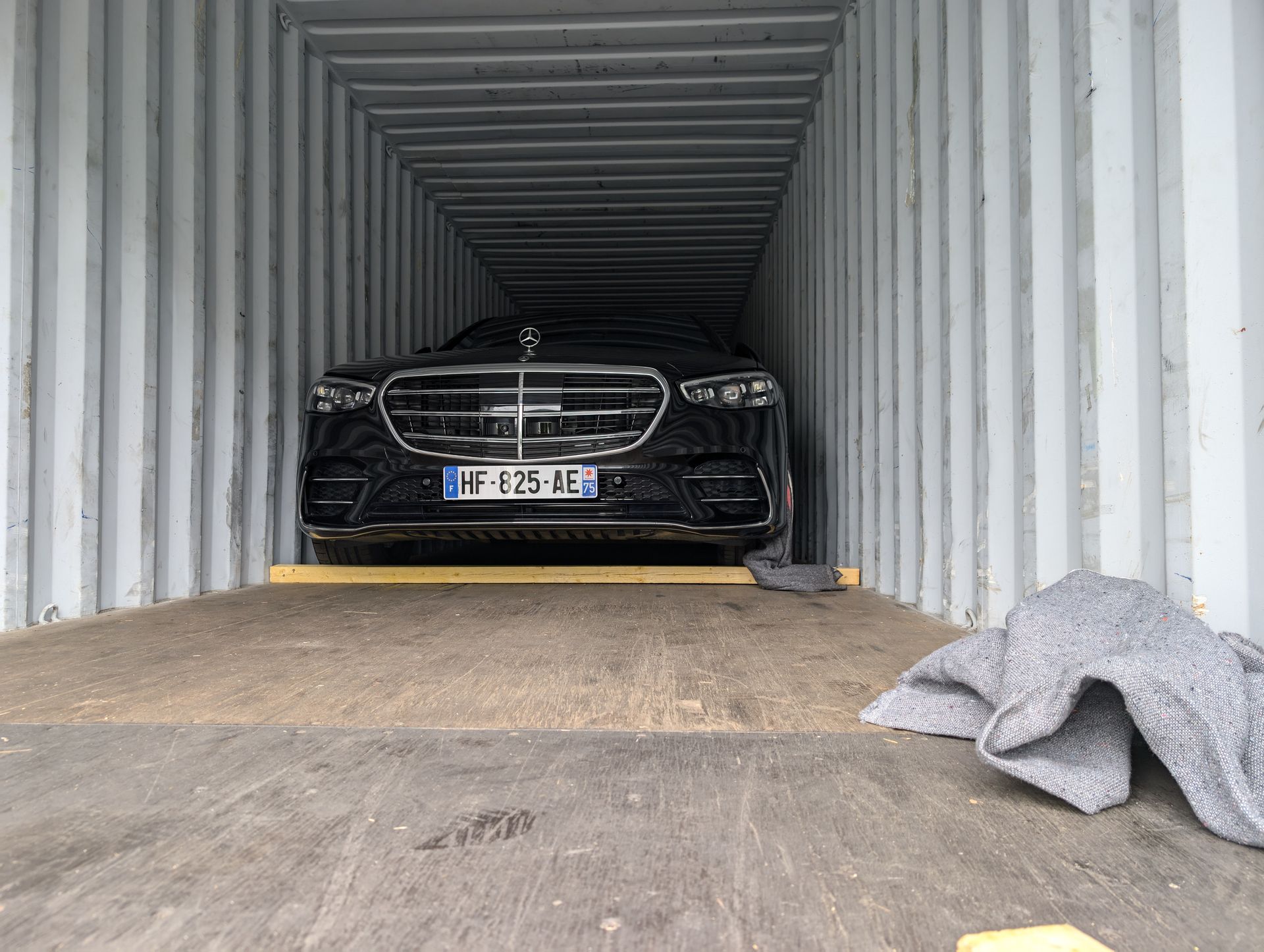 Une Mercedes noire est garée à l'intérieur d'un conteneur, avec une couverture grise sur le côté droit.