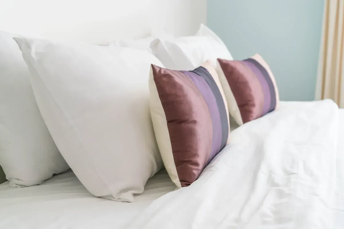 Cama blanca con almohadas; dos almohadas decorativas de color morado.