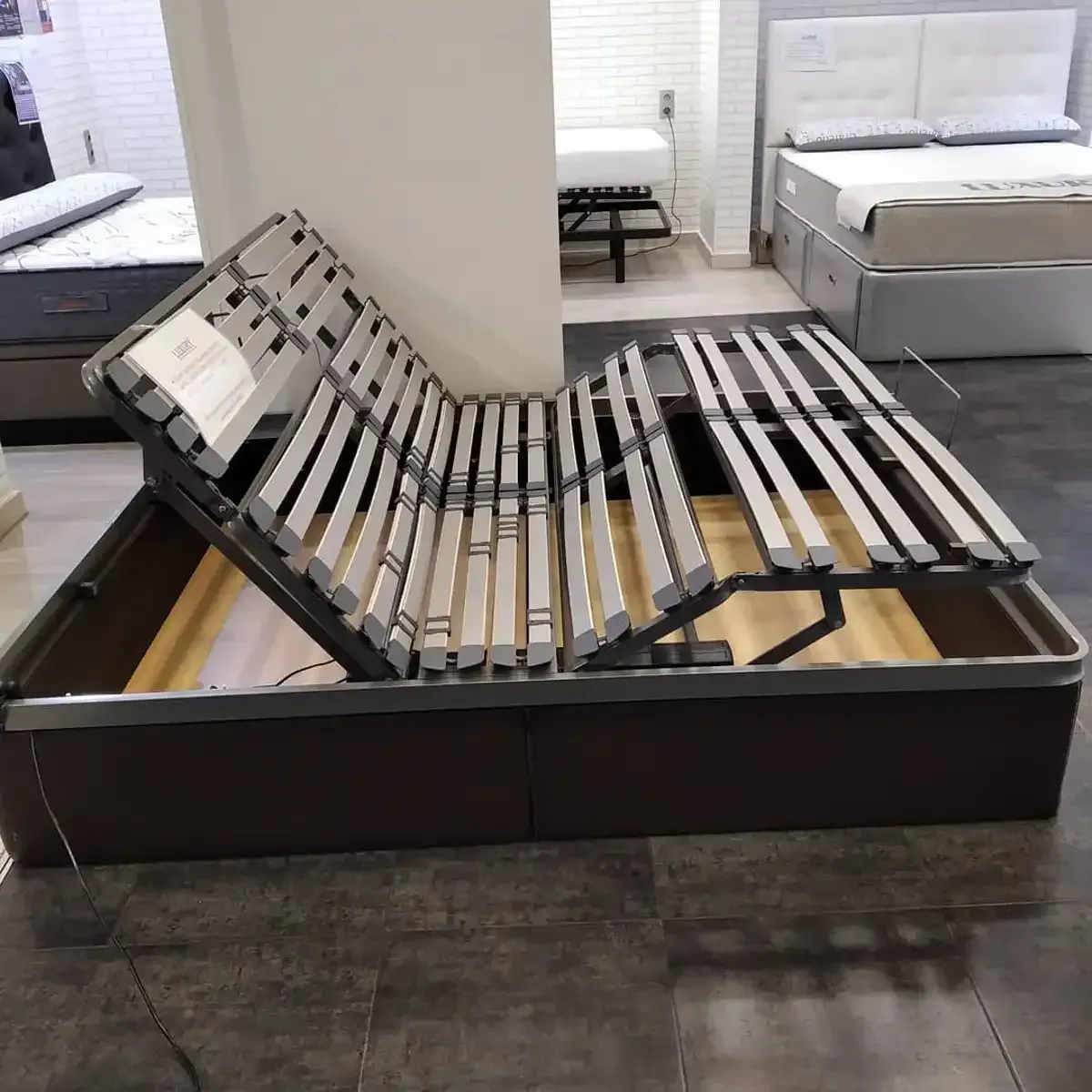 Estructura de cama ajustable, marrón, en un entorno de sala de exposición; secciones de cabecera y pies elevadas.